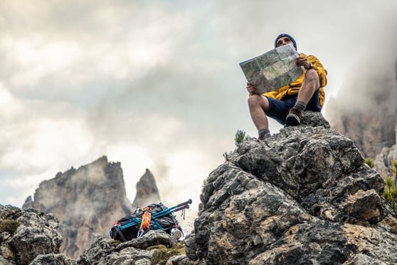 Young man hiker sitting on stone mountain reading map, with cloudy sky and fog. 