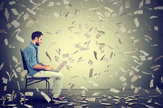 Man sitting with a laptop on his lap, while money falls around him.